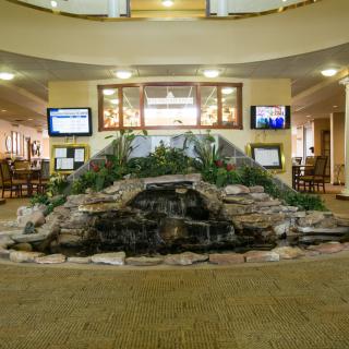 A spacious lobby area featuring a central fountain with surrounding seating and multiple screens.