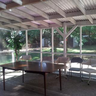 A sunlit room with a table and chairs, surrounded by large windows and greenery.