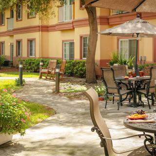 A peaceful outdoor patio area with tables, chairs, and umbrellas on a stone pathway, surrounded by greenery and adjacent to a building.