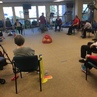 A group of seniors participating in a seated exercise class in a spacious indoor setting.