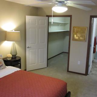 A cozy bedroom featuring a bed with red bedding, a bedside lamp, and an open closet door revealing shelving space.