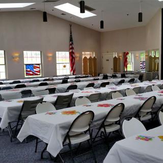 A banquet hall set up with tables and chairs covered with white tablecloths, featuring an American flag in the background.