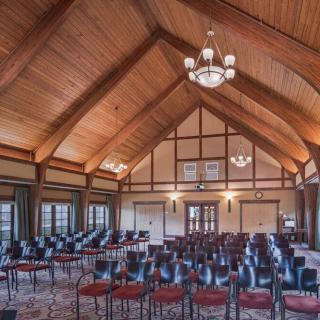 A spacious conference room with a wooden ceiling and rows of chairs arranged for an event, illuminated by elegant chandeliers.