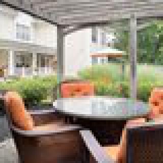 A cozy outdoor patio area featuring a round glass table surrounded by cushioned chairs under a pergola, with a view of the garden and house in the background.