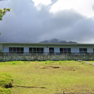 A single-story building with a stone wall and large windows, set against a cloudy sky and surrounded by grass.