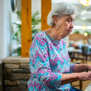 An elderly woman is playing the piano indoors, wearing a colorful shirt.