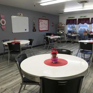 A well-organized classroom with round tables and chairs, featuring a whiteboard on the wall and windows allowing natural light.