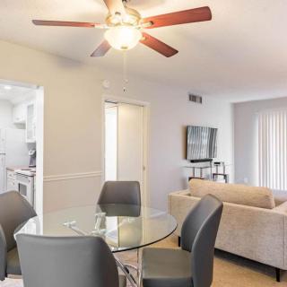 A modern apartment interior featuring an open-plan dining and living area with a ceiling fan, glass dining table, upholstered chairs, comfortable sofa, and mounted television.