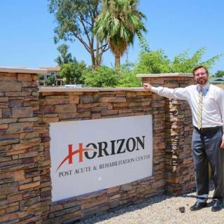A man stands beside a brick wall featuring a business sign for Horizon against a clear blue sky.