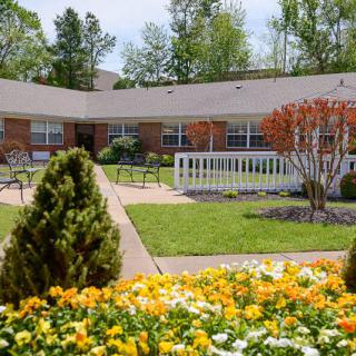 A beautiful garden with vibrant flowers in front of a brick building, surrounded by trees and benches.