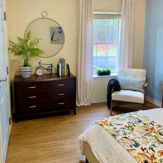A cozy bedroom with a wooden dresser, round mirror, and a comfortable chair by the window.