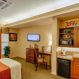 A well-lit hotel room with a desk, wall-mounted television, and kitchenette area.