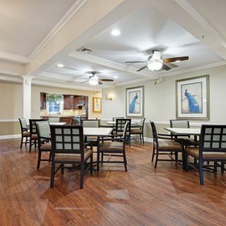 A spacious dining area with several tables and chairs, featuring wooden flooring, ceiling fans, and framed artwork on the walls.