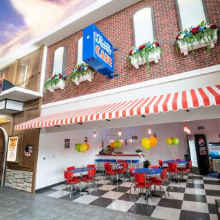 A vibrant diner with red chairs and a checkered floor, featuring a striped awning and flower boxes.