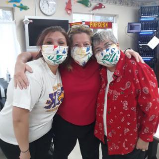 A joyful group of three women posing together, all wearing colorful masks, in a cheerful indoor setting.