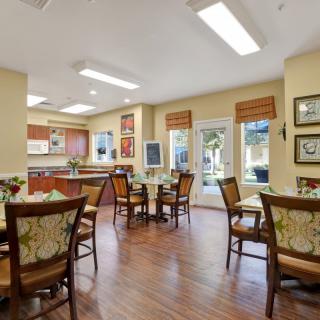 A cozy dining area with several tables and chairs, adjacent to a kitchen space, bathed in natural light from large windows.