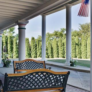 A serene porch with benches overlooking a manicured garden, featuring classic columns and a small flag hanging from the ceiling.