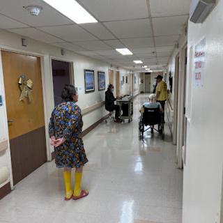A scene in a hospital hallway where a woman stands while a caregiver assists a patient in a wheelchair.