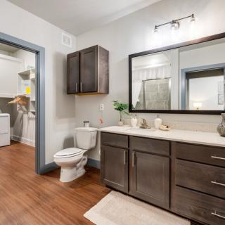 A modern bathroom with a large mirror, dark wood cabinetry, and hardwood flooring, adjacent to a laundry room.