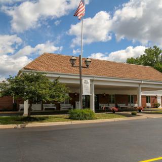 A front view of a hospital or clinic entrance with an American flag flying above, surrounded by trees and under a partly cloudy sky.