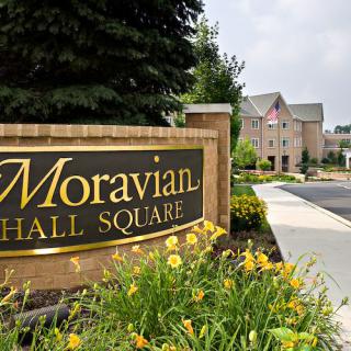 Entrance sign for Moravian Hall Square with a pathway and building in the background surrounded by flowers.