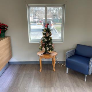 A small Christmas tree is displayed on a table next to a chair in a cozy room with a window.