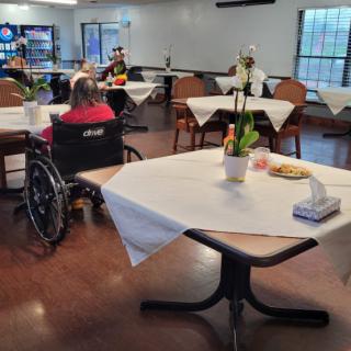 A nursing home dining area with tables covered in white tablecloths and a person in a wheelchair.