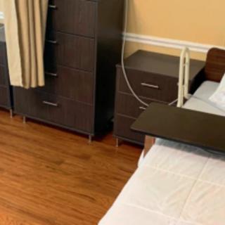 A room featuring a hospital bed with adjacent furniture, including a nightstand and dresser, set on a hardwood floor.