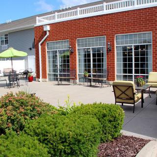 A cozy outdoor patio area with seating and an umbrella, adjacent to a building with large windows and surrounded by greenery.
