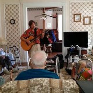 A musician plays guitar for an audience in a cozy living room setting.