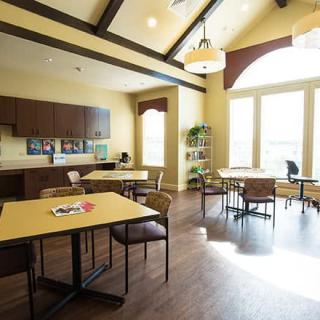 A well-lit dining area with tables and chairs, adjacent to a kitchen space with large windows allowing natural light.