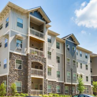 A modern multi-story apartment building with balconies and a stone facade, set against a partly cloudy sky.