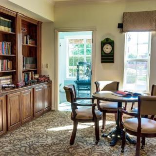 A cozy library room featuring a bookshelf, a round table with chairs, and sunlight streaming through the windows.