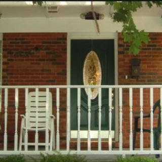 A porch with white railing, featuring a green door and several chairs in front of a brick house.