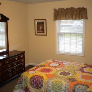 A cozy bedroom featuring a colorful quilt on the bed, a wooden dresser with a mirror, and a window with curtains.