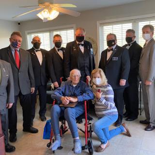 A formal gathering with an elderly individual in a wheelchair, surrounded by men in suits and women, all wearing masks.