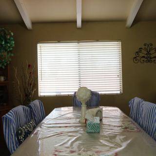 A dining room with a table covered in a plastic tablecloth and chairs with striped covers, illuminated by light filtering through window blinds.