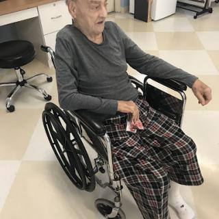 An elderly individual sitting in a wheelchair, dressed comfortably in plaid pants.