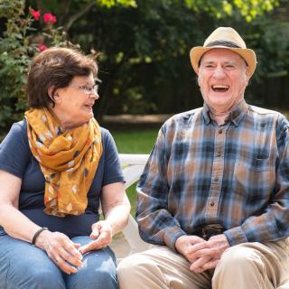 An elderly couple enjoying a sunny day in the garden, sharing laughter while sitting on a bench.