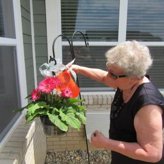 A senior woman waters flowers in a hanging pot outdoors.