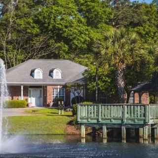 A cozy brick house with a dock overlooking a pond and fountain surrounded by lush greenery.