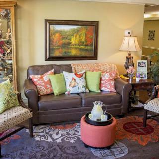 A cozy living room with a brown sofa adorned with colorful pillows, a landscape painting on the wall, and a decorative lamp on an end table.