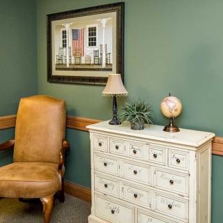 A cozy room corner featuring a leather chair, a vintage dresser with a lamp and globe, and a framed painting on the green wall.