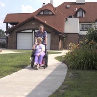 A caretaker pushes an elderly person in a wheelchair along a pathway outside a house.