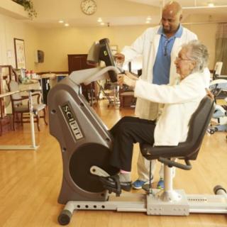 An elderly person receiving guidance from a trainer while using an exercise bike in a rehabilitation center.