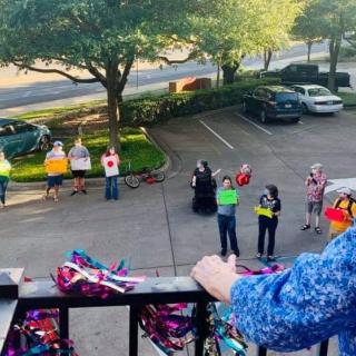A group of people stand in a parking lot, some holding signs and balloons, viewed from a balcony.