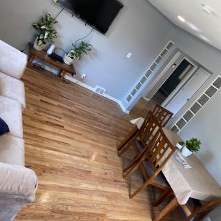 A cozy living and dining space with wooden flooring, featuring a sofa, dining table with chairs, indoor plants, and a wall-mounted TV.