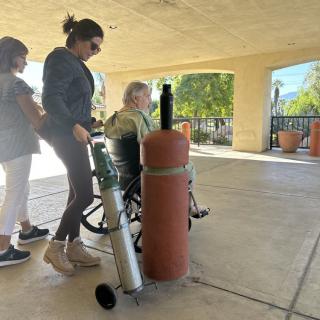 A woman in a wheelchair with an oxygen tank being assisted outside by two caregivers.