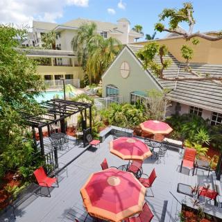 An inviting outdoor patio area with red umbrellas and seating, surrounded by lush greenery and a view of nearby buildings.