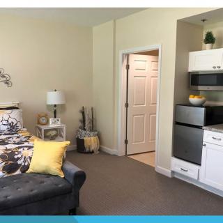 A cozy room featuring a sofa with floral and yellow pillows, a lamp on a side table, and a kitchenette in the corner.
